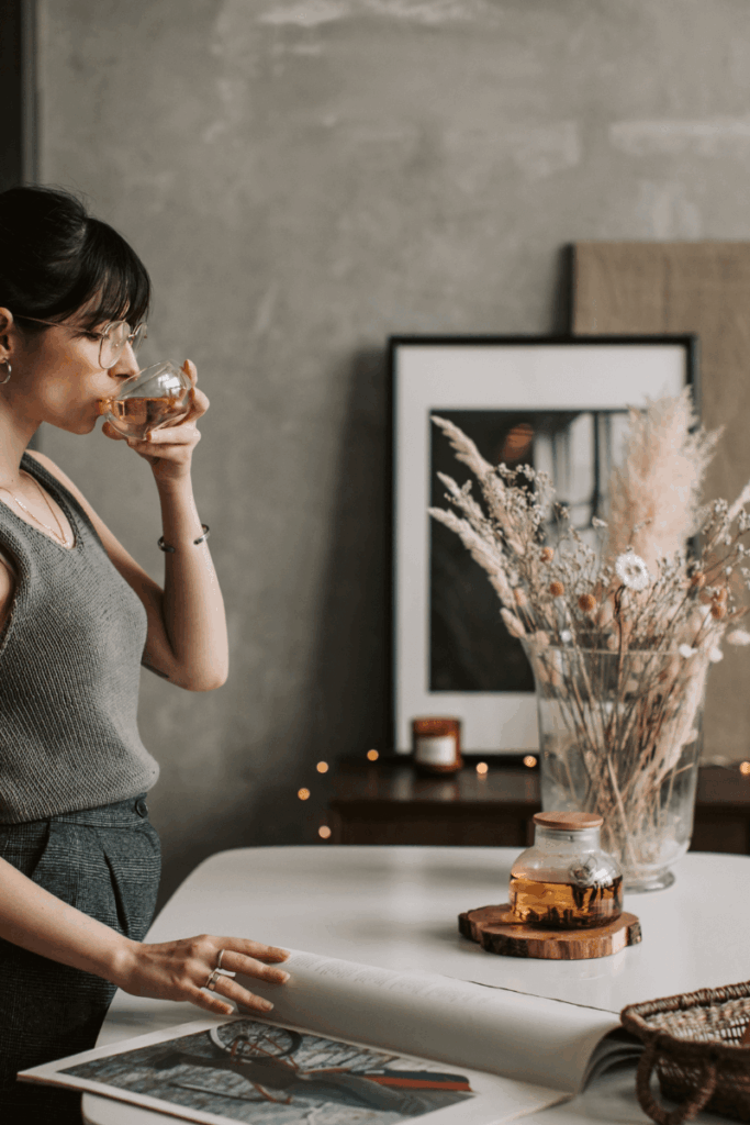 A woman standing near a table, sipping on her drink, while reading through a book that is lying on the table.
