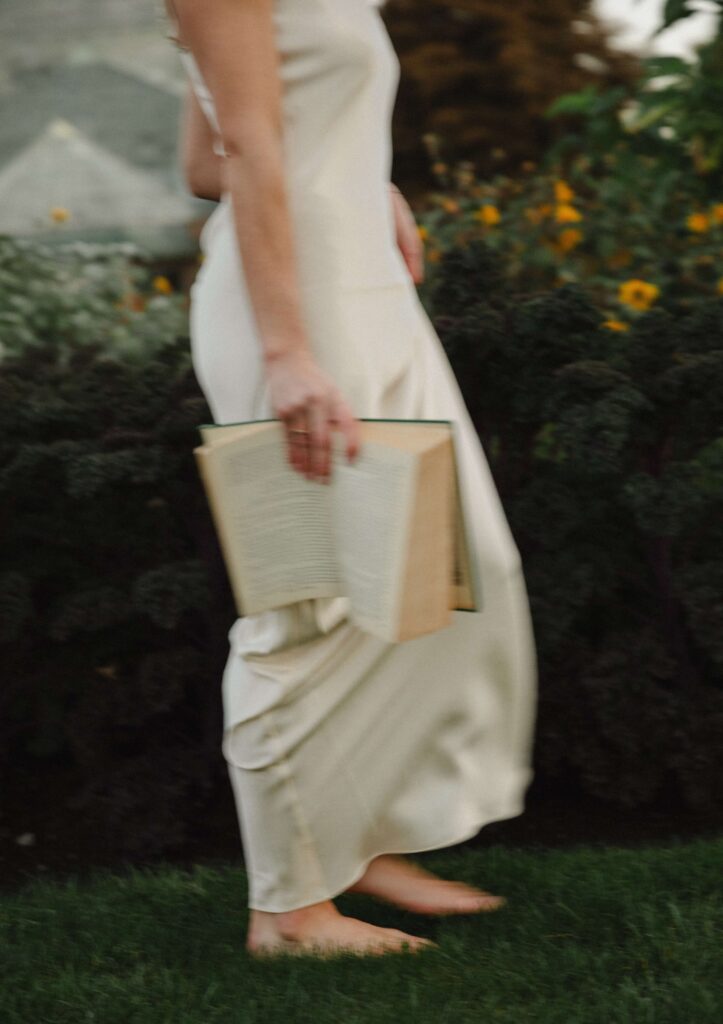 A woman standing barefoot on grass with a book in her hand, by her side. 