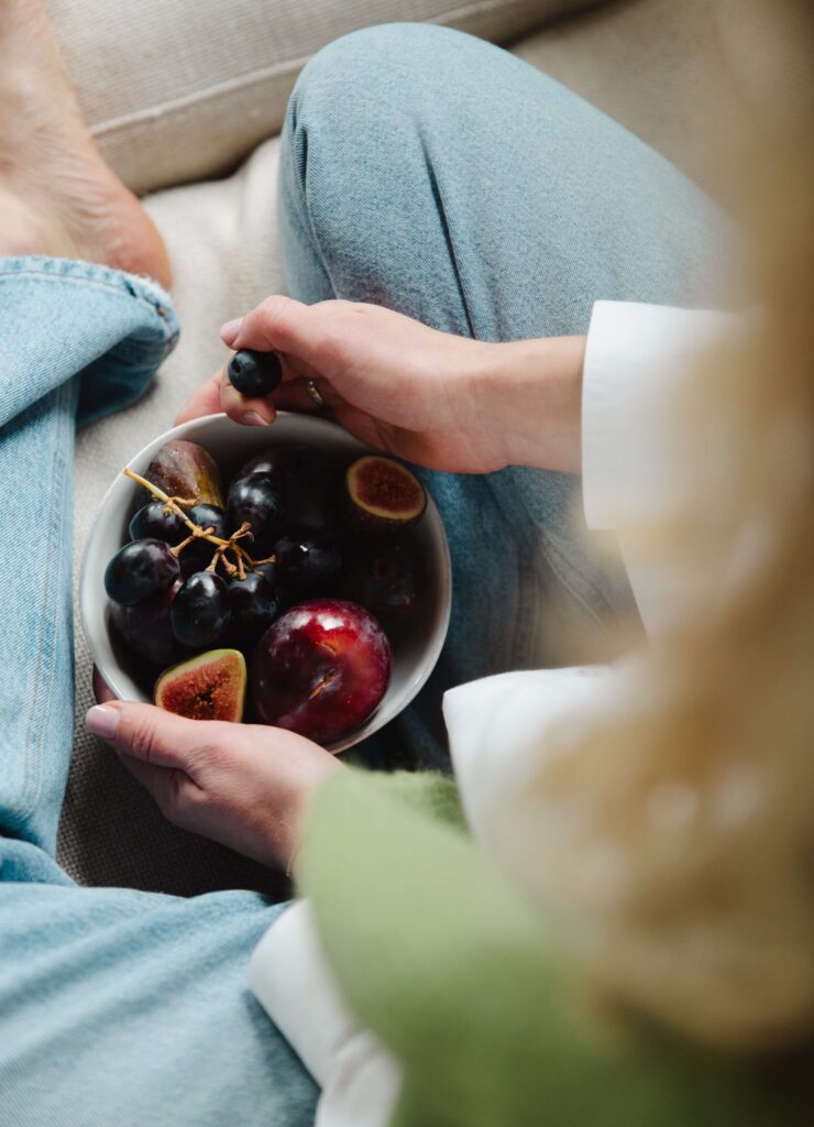 A woman in jeans, sitting on a couch, with a bowl of fruits. 