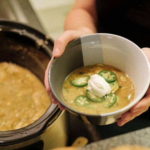 Bowl of green chicken chili soup topped with sour cream and sliced jalapeños, served fresh from a pot