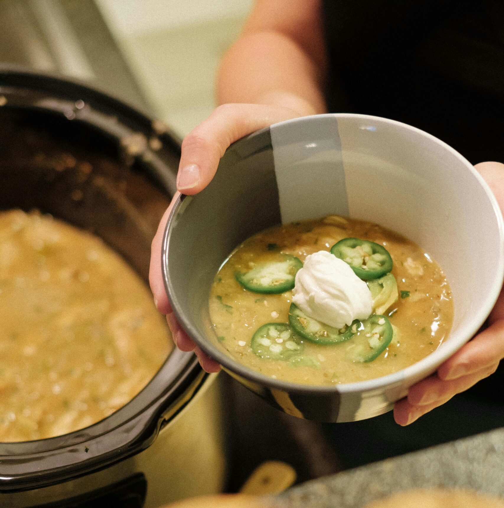 Bowl of green chicken chili soup topped with sour cream and sliced jalapeños, served fresh from a pot