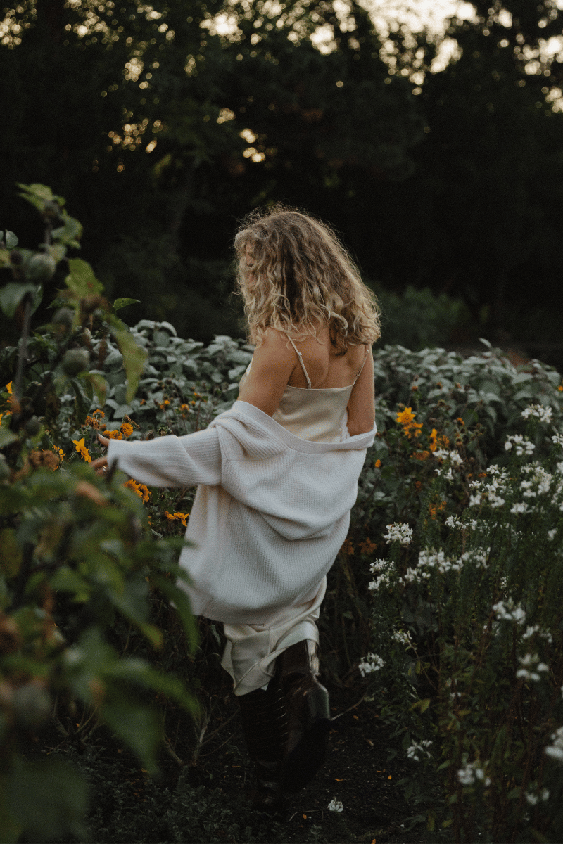Woman with curly blonde hair walking through a flower garden, wearing a cream dress and a white shawl surrounded by wildflowers.