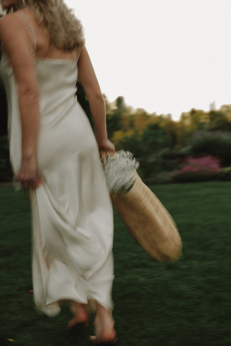 Woman in a flowing white dress walking barefoot through a garden, carrying a woven basket filled with flowers.
